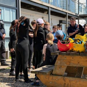 Group of people in black wetsuits gathered on a dock beside a row of colorful kayaks, ready for a water activity instructor nearby.