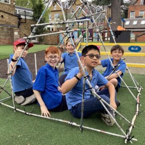 Five children in blue shirts pose inside a geometric frame made of metal poles on a playground, smiling at the camera.