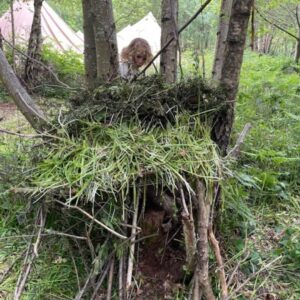 Child peeking from a leafy, nest-like fort built around a tree in a forest campsite, with tents in the background.