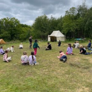 Group of children seated in a circle on a grassy field at a campsite, with tents and a beige canopy tent in the background.