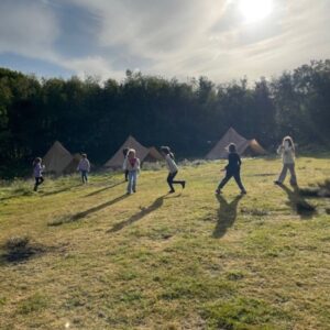 A group of people playing tag or running across a sunlit camping field with tents set up along the tree line in the background.