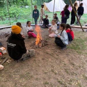 Children sit in a circle around a small campfire under a canopy, with tents visible in a wooded campsite behind them.