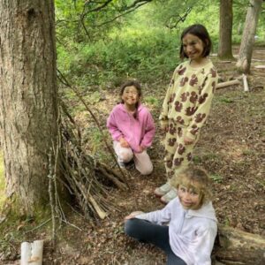 Three kids in a forest posing and smiling near a pile of sticks and logs, dressed in casual clothes for outdoor play.