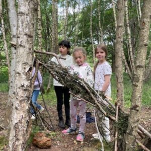 Three children in a birch forest constructing a shelter from branches and twigs on a dirt path.