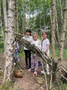 Three children in a birch forest building a makeshift shelter from sticks and branches.