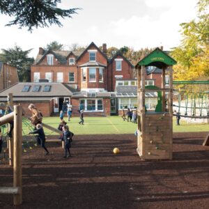 Children playing on a wooden school playground with climbing nets and slides, brick houses in the background.