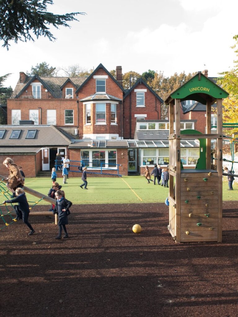 Children playing on a wooden school playground with climbing nets and slides, brick houses in the background.