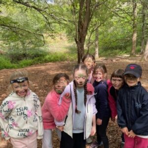 Group of smiling kids in a forest, posing together on a leaf-covered path. Some have face paint and braided hair, wearing jackets and sneakers.