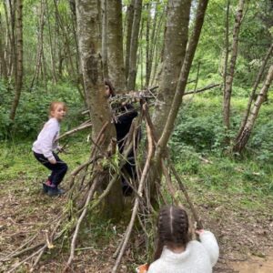 Children building a shelter from sticks and branches between trees in a forest setting behind them