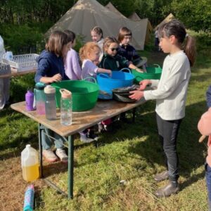 Kids at a campsite washing dishes in large plastic tubs on a wooden table, with tents and trees in the background.