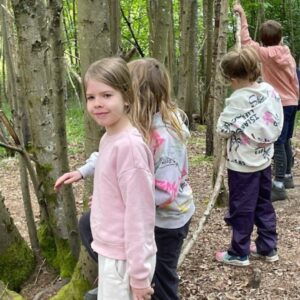 Four children in pastel clothing stand in a forest, touching a row of sticks laid on the ground between trees as they explore.