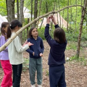Four kids in a forest campsite tying a branch to form a shelter, tents visible in the background.