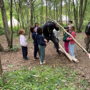 Group of children in a forest watching a man walk along a long, curved wooden log as a balance obstacle course stunt