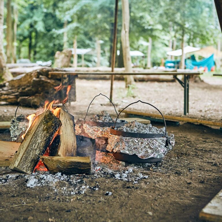 Campfire with burning logs and two foil-wrapped pots cooking over the flames at a forest campsite.