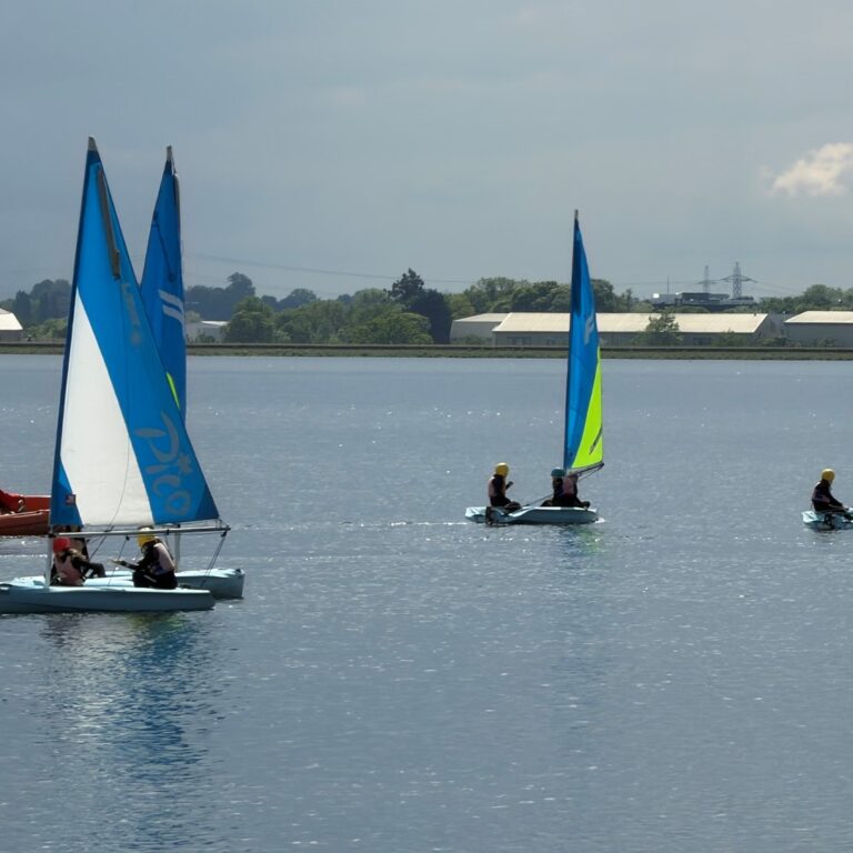 Several small sailboats glide on a calm lake, blue and white sails with two riders each, wearing life jackets; a red motorboat is nearby, shoreline in the distance.