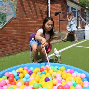 Girl sitting on a chair, fishing colorful balls from a blue ball pit in a sunny outdoor playground by a brick wall.